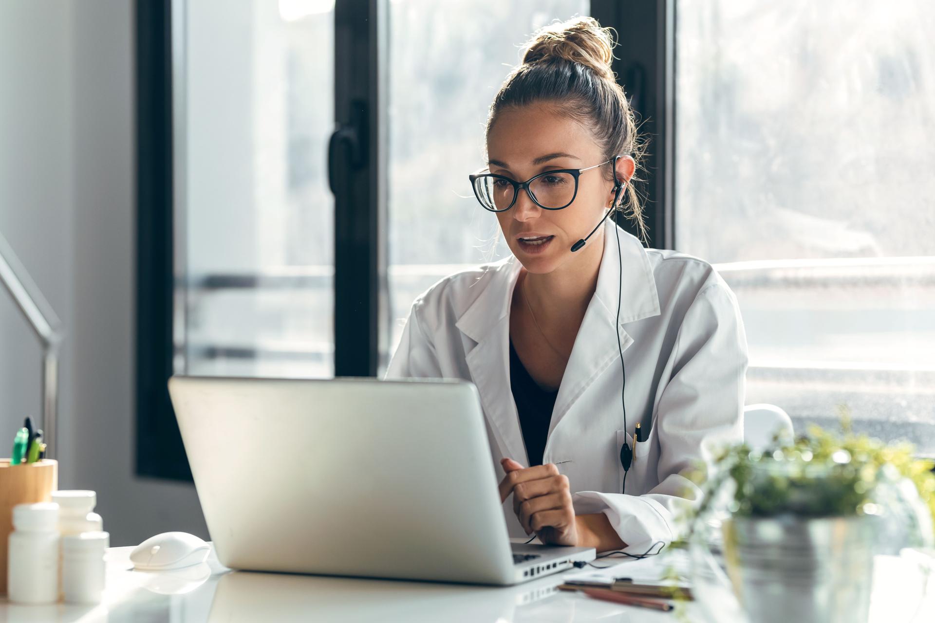 Female doctor talking while explaining medical treatment to patient through a video call with laptop and earphones in the consultation.