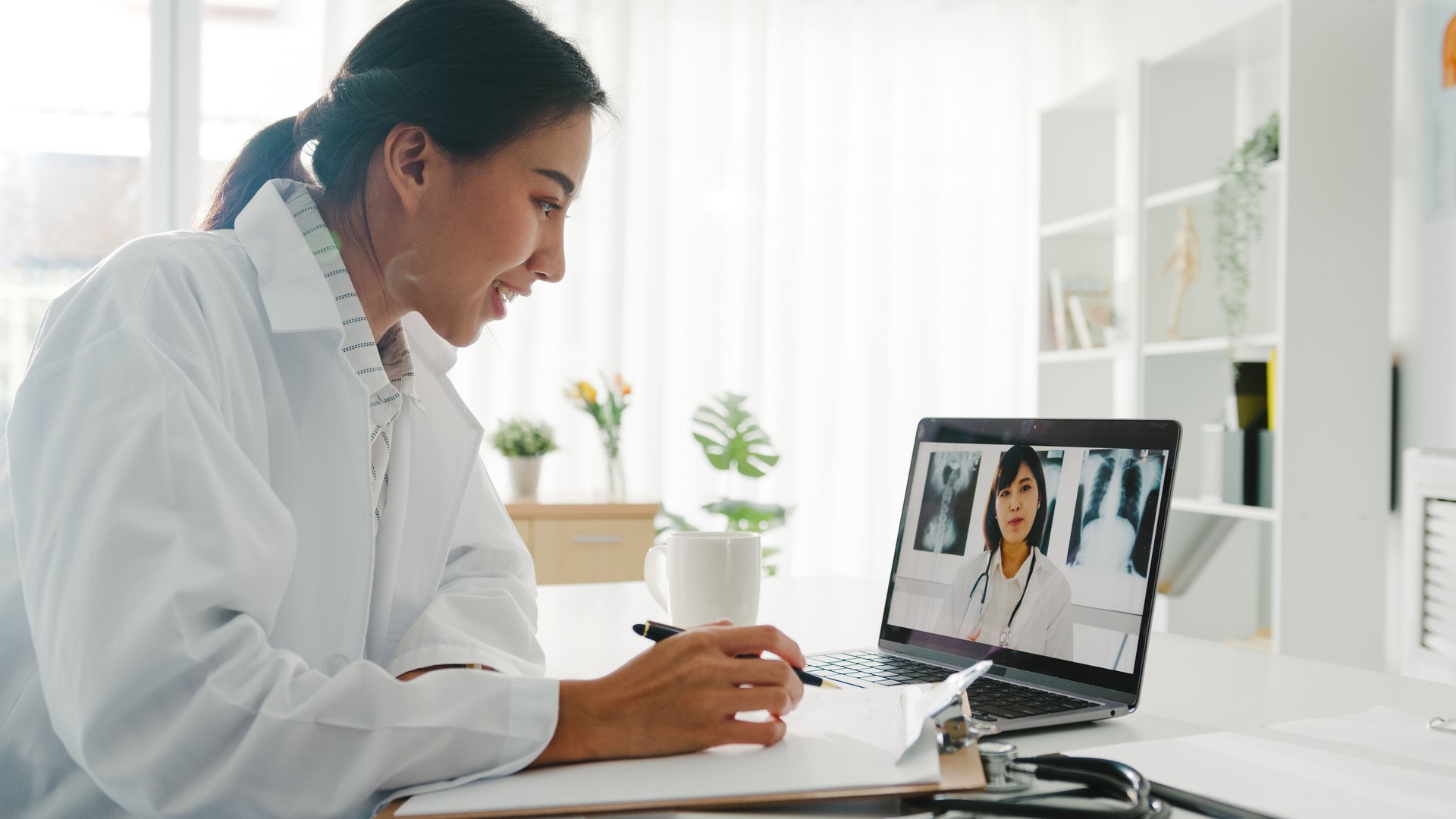 Young Asia lady doctor in white medical uniform using laptop talking video conference call with senior doctor at desk in health clinic.